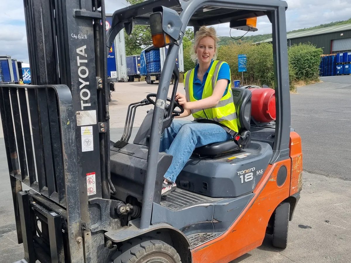 Young woman wearing a high-visibility vest seated at the controls of a forklift in a warehouse yard.